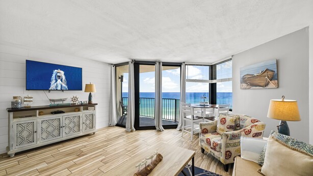 Un salón amplio con piso de madera, un televisor grande, un comedor con mesa y sillas, y vista al océano a través de amplias ventanas.