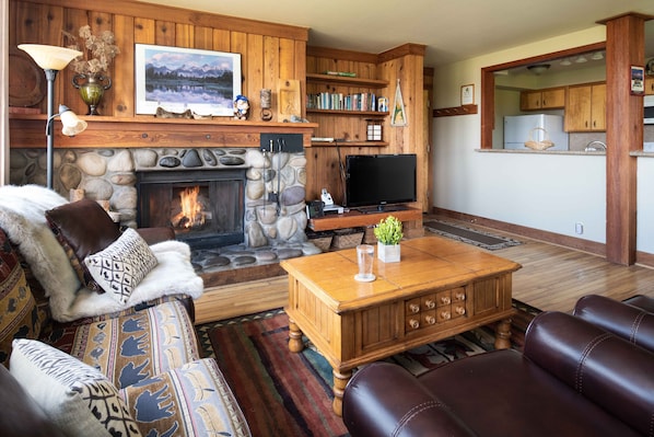 A cozy living room with a stone fireplace, a flat-screen TV, leather sofas, and a wooden coffee table. - Outpost: Nez Perce A3 - Hot Tub Access! (Teton Village)