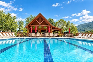 Une piscine avec des chaises longues et des parasols, un bâtiment en bois doté d’un toit triangulaire, et un paysage montagneux en arrière-plan.