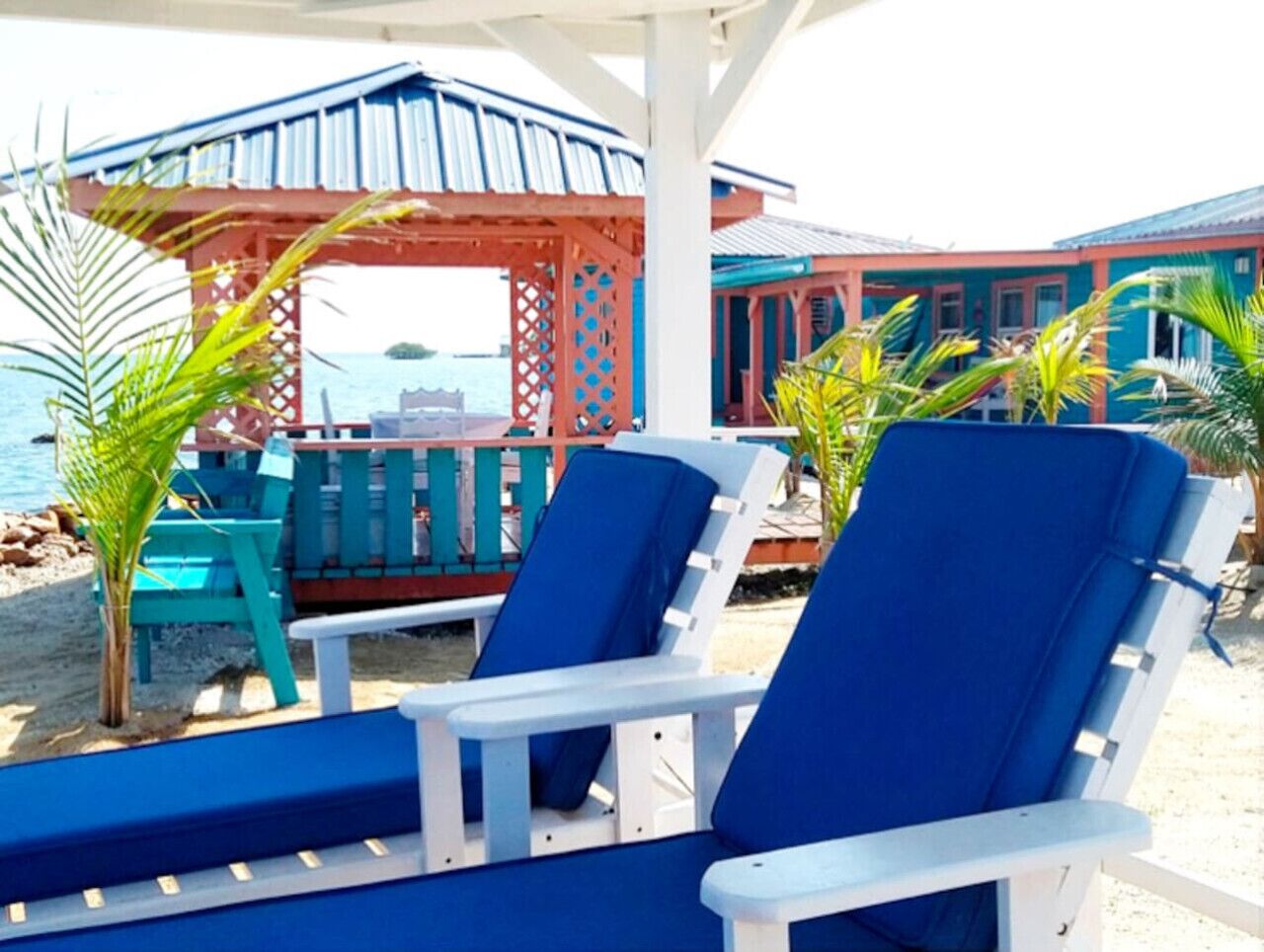 A beachside lounge area with blue and white lounge chairs, a wooden gazebo, and a view of the sea.