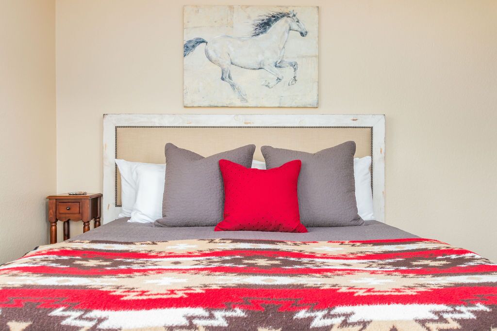 A bed with a patterned blanket, a white headboard, and three pillows in white, gray, and red.