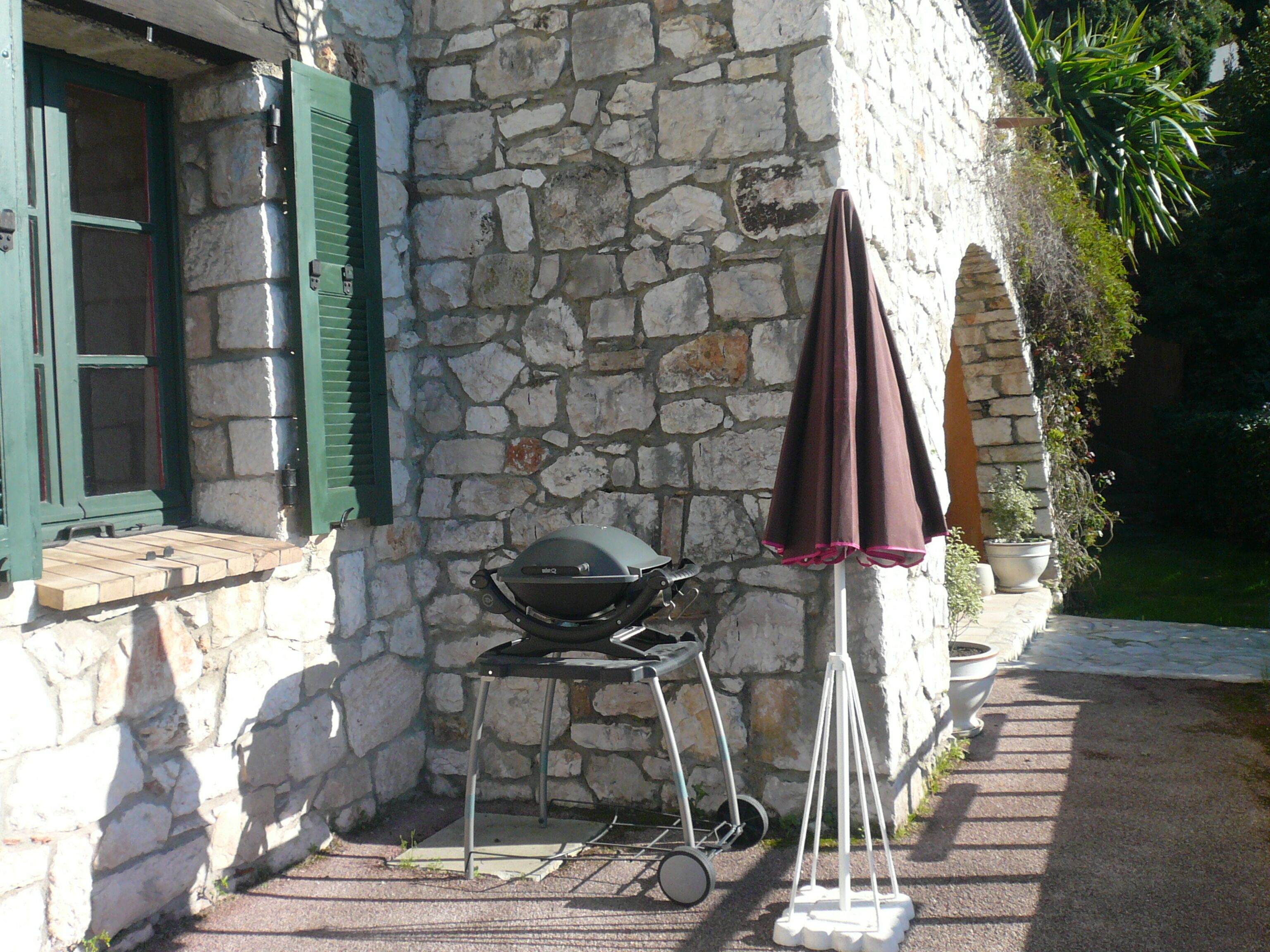 Un bâtiment en pierre avec un barbecue à l’extérieur, une fenêtre avec des volets verts et un pot de fleurs.