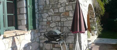 Un bâtiment en pierre avec un barbecue à l’extérieur, une fenêtre avec des volets verts et un pot de fleurs.