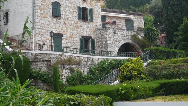 Un edificio de piedra con contraventanas verdes, un balcón y una escalera que lleva a la entrada.