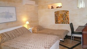 Una habitación con dos camas, un escritorio, una silla, un equipo de aire acondicionado, un cuadro enmarcado y una ventana de vitral.