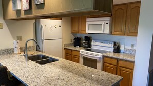 Une cuisine dotée de plans de travail en granit, d’un évier en acier inoxydable, d’un réfrigérateur blanc, d’un four à micro-ondes, d’une cuisinière, de meubles de rangement en bois et d’une machine à café.