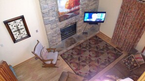 Un salon avec une cheminée en pierre, un téléviseur fixé au mur, un tapis à motifs et un fauteuil à bascule en bois.