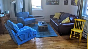 Un salon avec un canapé bleu, un canapé en cuir marron, un fauteuil jaune, une petite table et une fenêtre ornée d’œuvres d’art sur le mur.