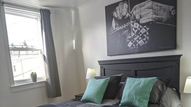 Une chambre à coucher avec un lit, une table de chevet, une lampe, une fenêtre avec des rideaux et un tableau au mur.