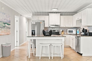 Una cocina con muebles blancos, una isla central con taburetes, un refrigerador, un microondas y una cafetera.