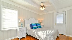 A bedroom with a white bed, two bedside tables with lamps, a ceiling fan, and a window with blinds.