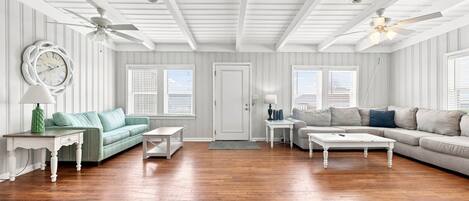 A spacious living room with hardwood flooring, a green sofa, a grey sofa, two white coffee tables, and a white door.