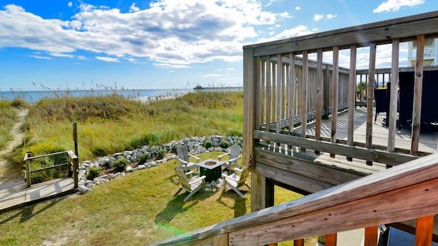 A wooden deck overlooking a beach with a grassy area and a small table with chairs.
