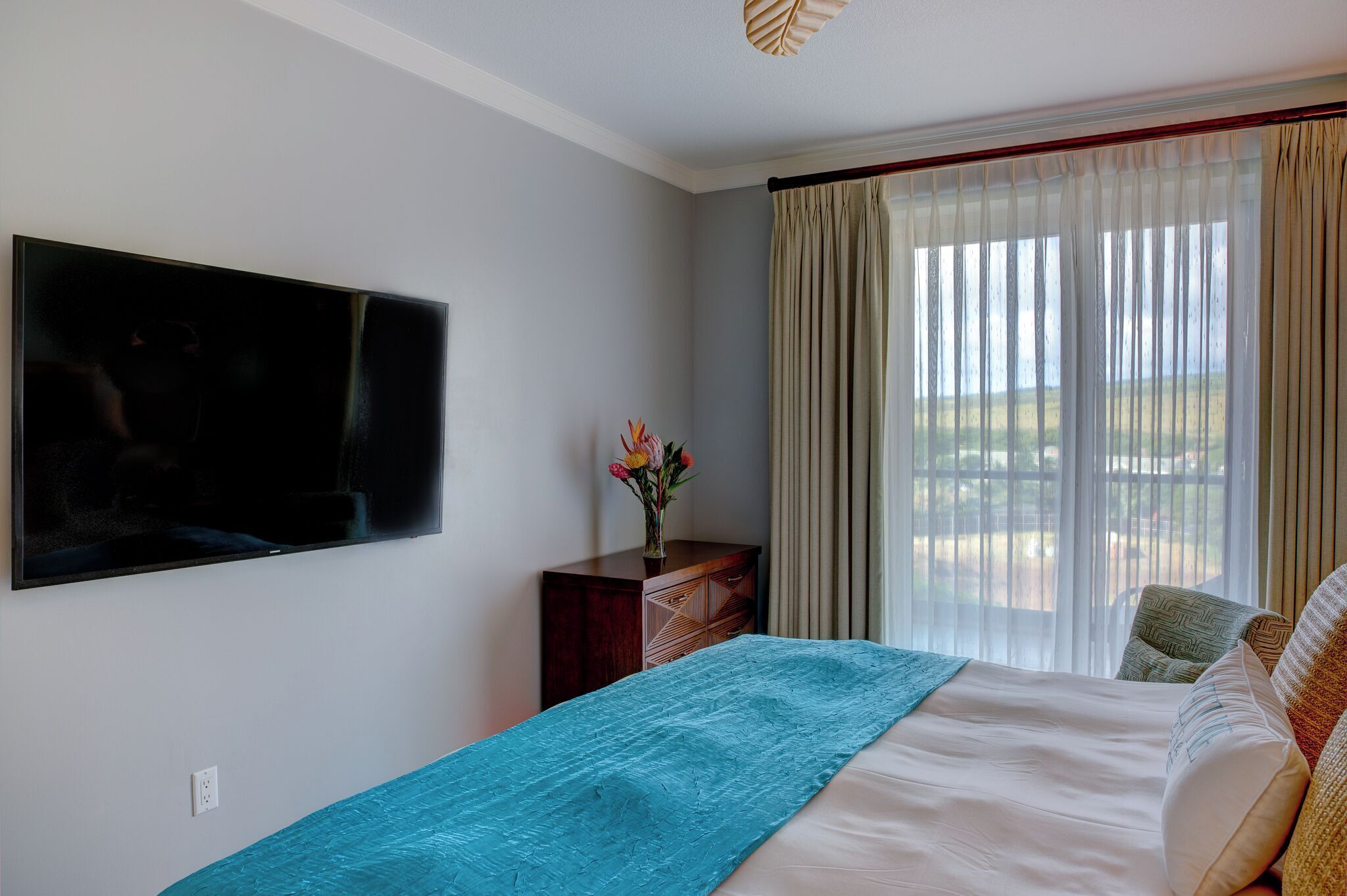 A bedroom with a bed, a TV mounted on the wall, a dresser, and a window with curtains.