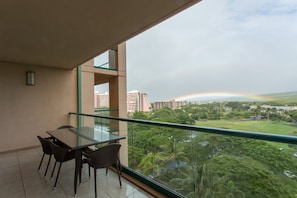 Un balcón con una mesa y sillas de vidrio, con vistas a un paisaje urbano con edificios y un arcoíris.