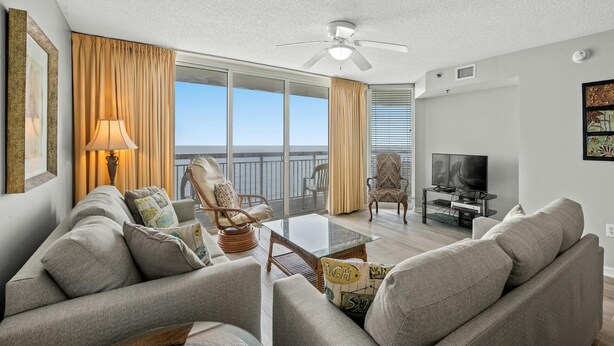 A spacious living room with a balcony, a ceiling fan, and a television. - Charming 15th Floor Condo, Indoor and Outdoor Pool | Crescent Shores - S 1508 (North Myrtle Beach)