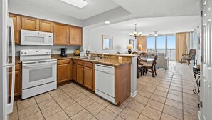 A kitchen with wooden cabinets, a microwave, stove, and dishwasher. There is a dining area with a table and chairs, and a view of the ocean through large windows. - Charming 15th Floor Condo, Indoor and Outdoor Pool | Crescent Shores - S 1508 (North Myrtle Beach)