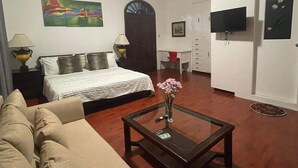 Ein Wohnzimmer mit Sofa, Couchtisch mit Vase, Bett mit weißer Decke, Fernseher an der Wand und Tür mit Bilderrahmen.