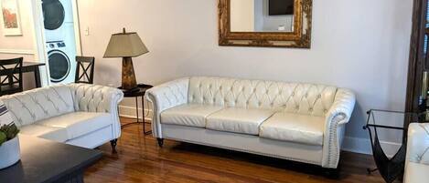 A living room with a white leather sofa, a wooden floor, a lamp, and a large mirror on the wall.