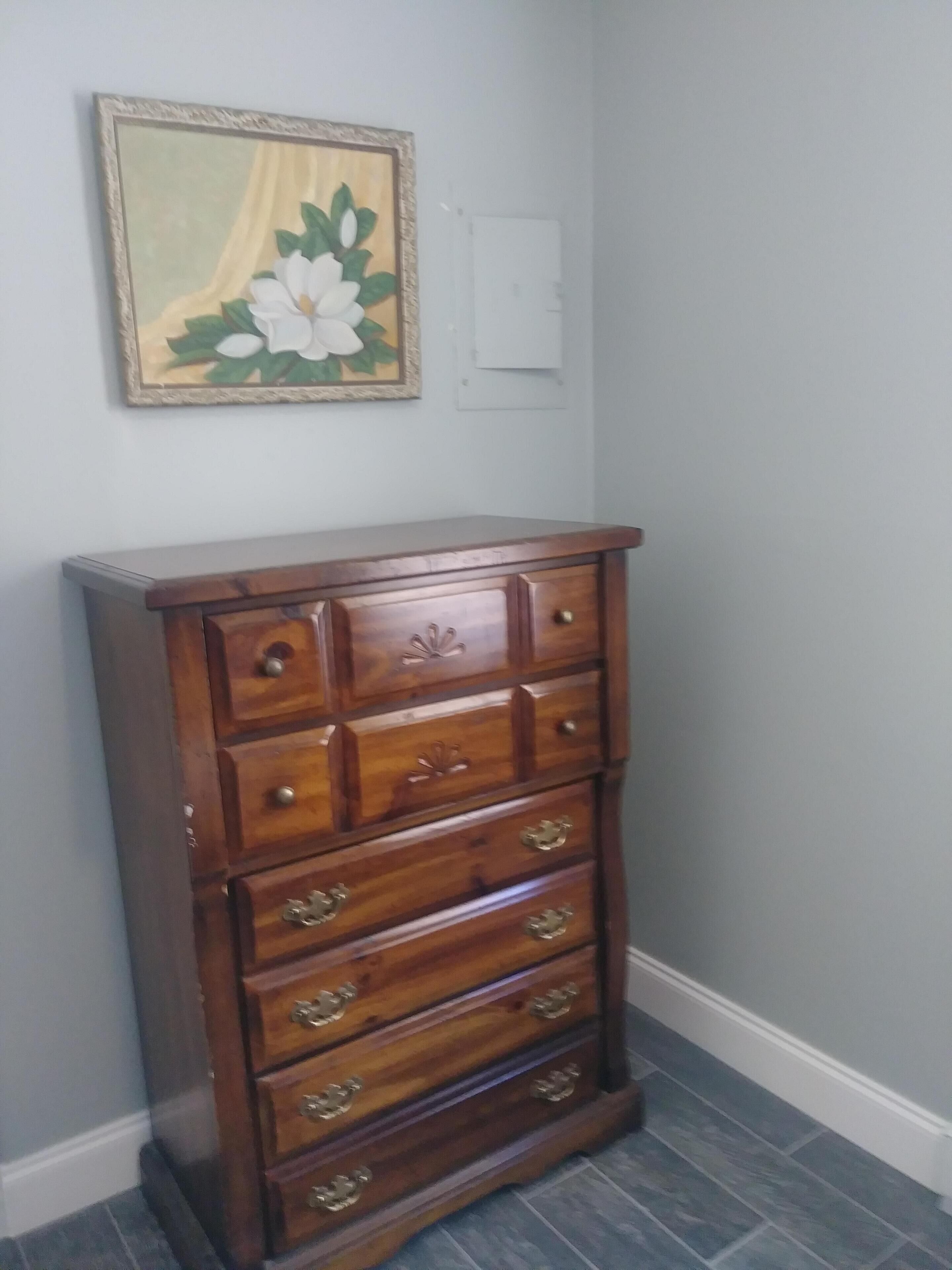A wooden dresser with multiple drawers and brass handles, positioned against a wall with a framed floral painting above it.