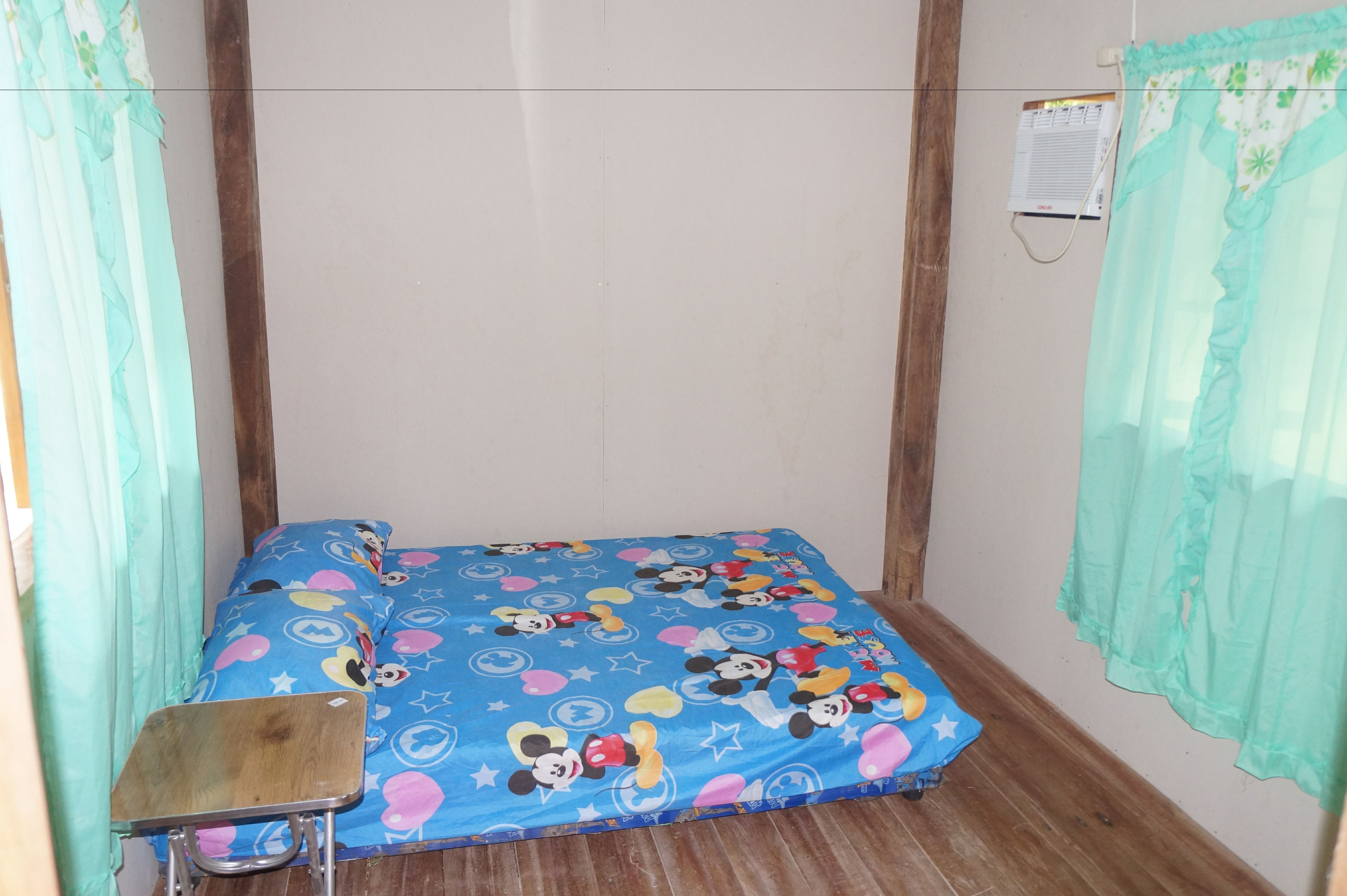 A small, single-bed room with a blue Mickey Mouse themed bedspread, a wooden desk, and a window with a green curtain.