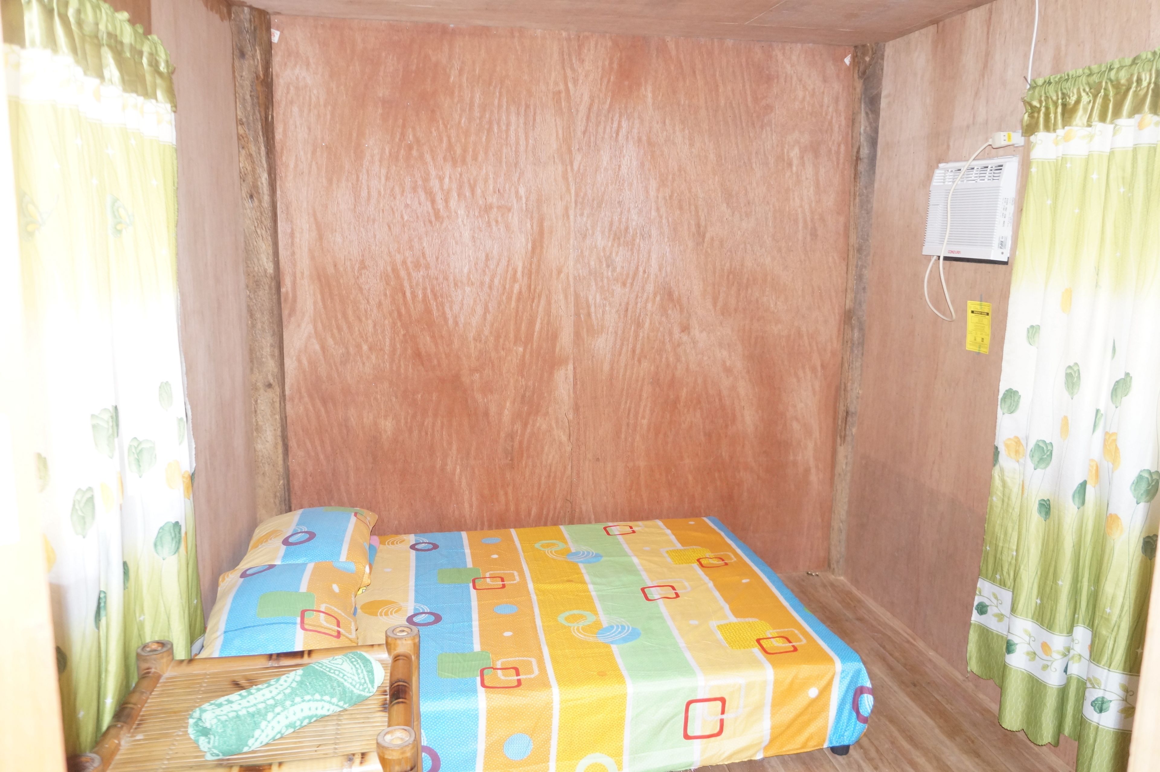 Une petite chambre avec un lit simple, une couvre-lit aux couleurs vives, un tapis en bambou et un climatiseur fixé au mur.
