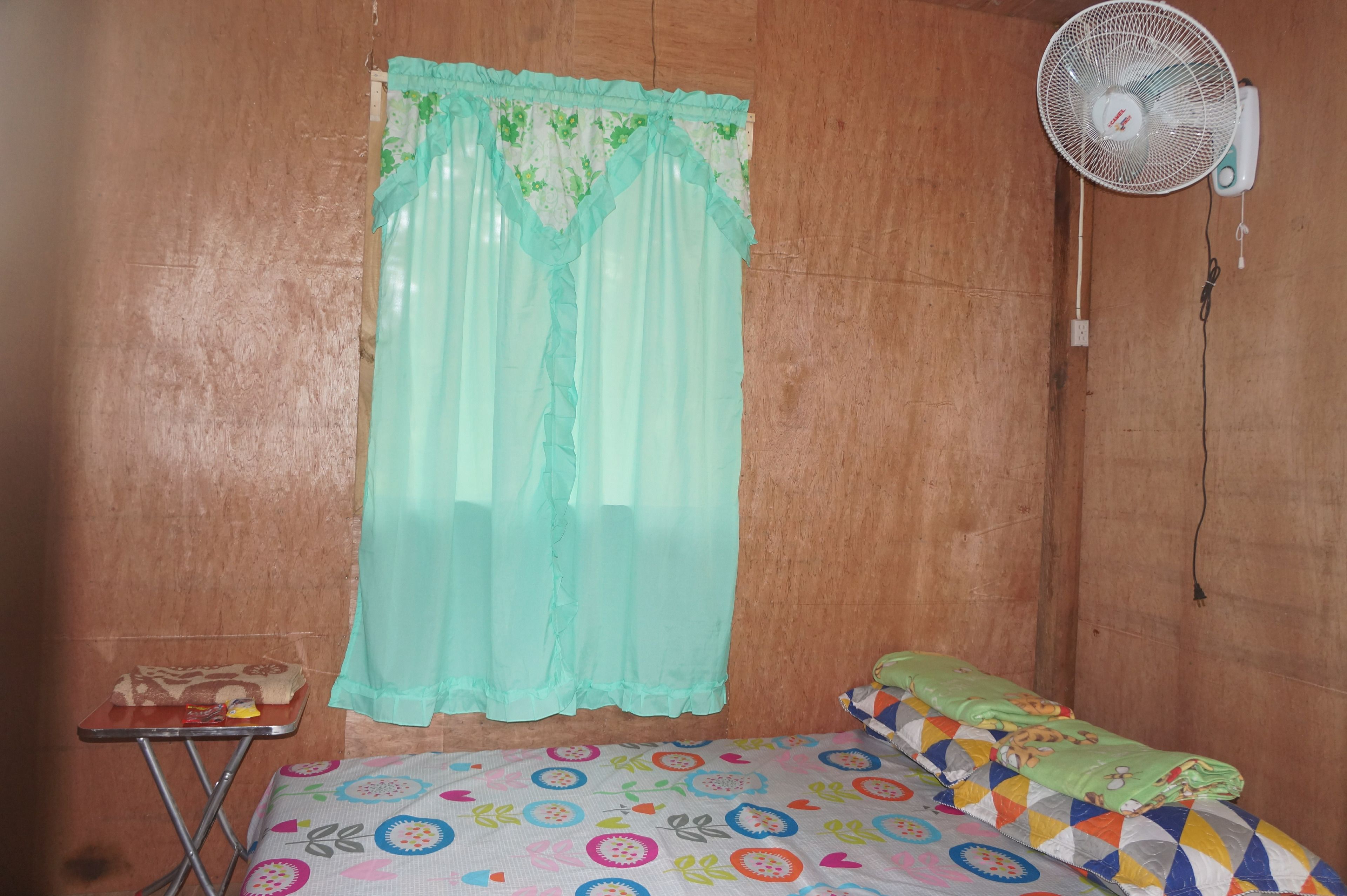 A small room with a bed, a table, a fan, and a curtain.
