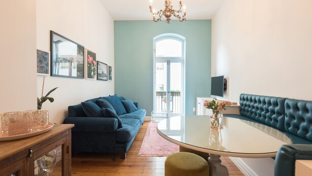 A living room with a dark blue sofa, a glass coffee table, a round table, and a blue upholstered bench. There are framed pictures on the wall, a chandelier, and a TV mounted on the wall. - The Designer's Residence (Auckland)