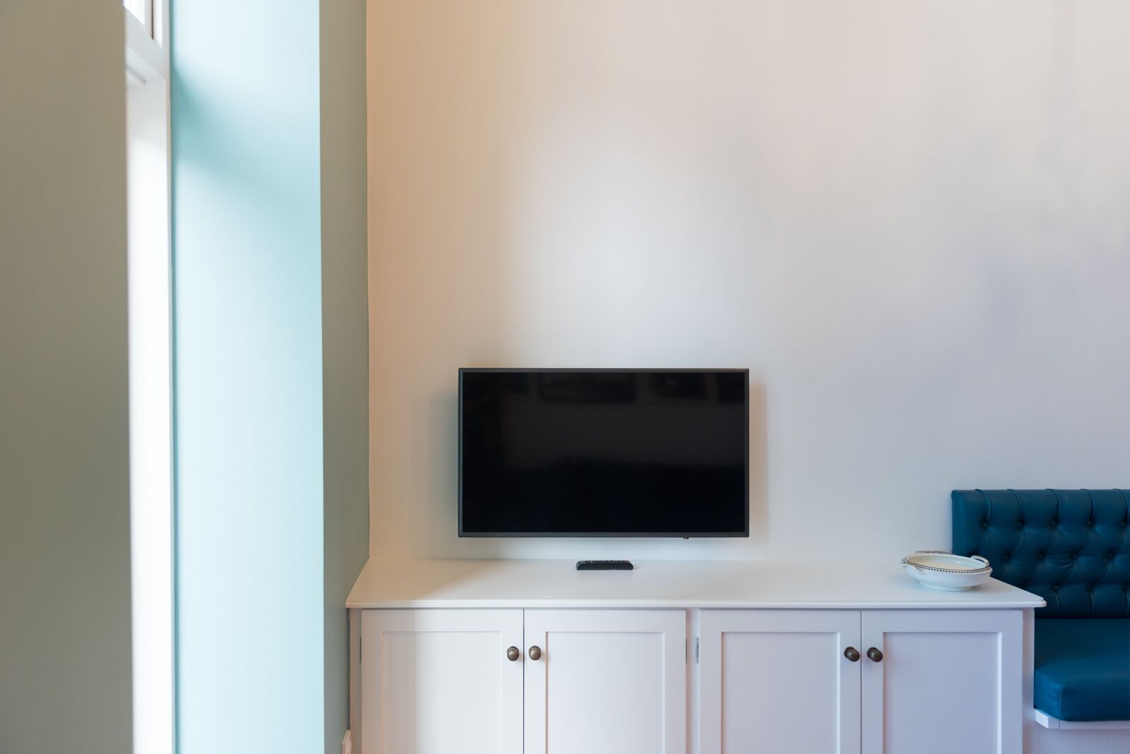 A white cabinet with a flat-screen TV mounted above it, a teal upholstered chair, and a white door with a handle.