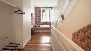 A narrow hallway with a wooden floor, a bed with a floral-patterned headboard, and a window with blinds. - The Designer's Residence (Auckland)