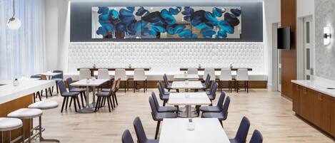 Un comedor moderno con mesas blancas y sillas azules, un cuadro abstracto grande en la pared y una encimera de madera con un televisor.