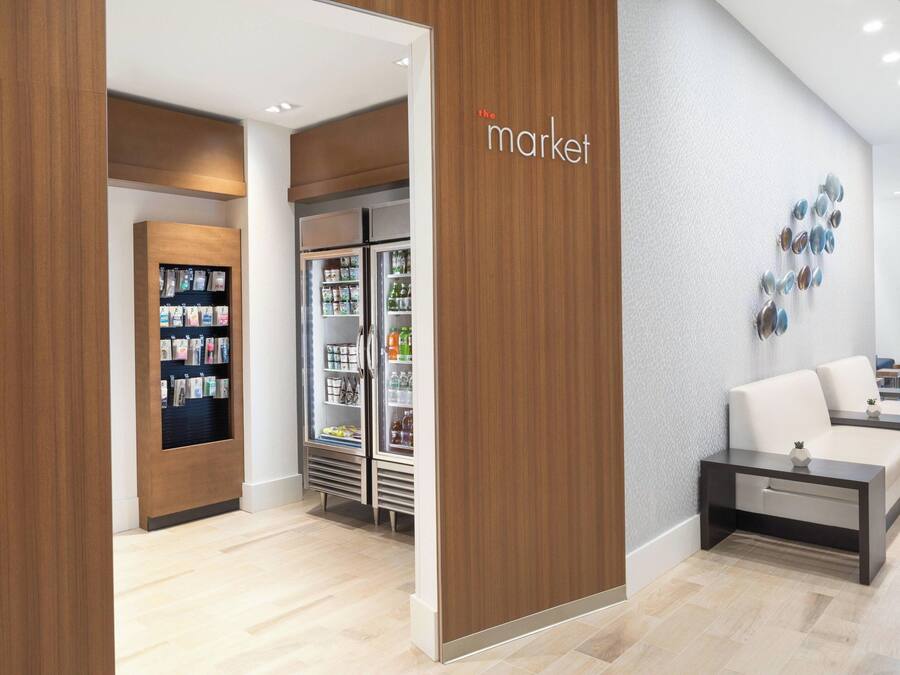 A modern hotel lobby with a wooden reception desk, a seating area with sofas, and a refrigerator stocked with beverages.