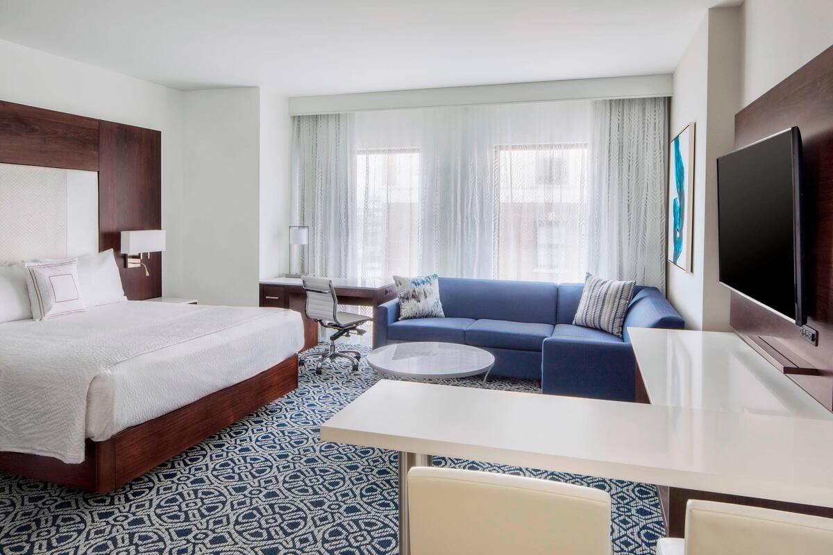 A hotel room with a large bed, a sofa, a desk, a TV, and a patterned carpet.