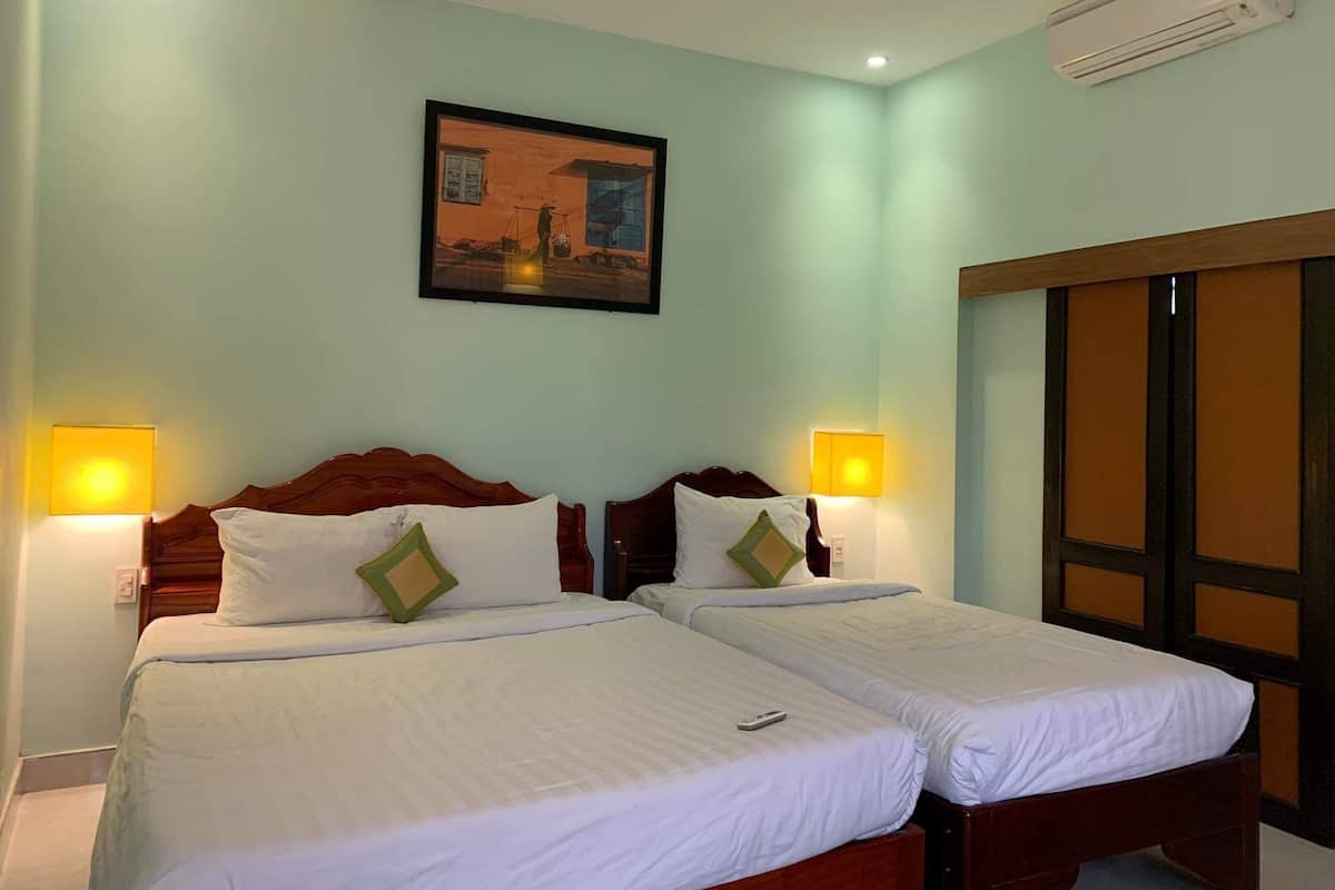 A hotel room with two beds, each with white bedding and green pillows. There are wall-mounted lamps on either side of the beds. A framed picture hangs on the wall above the beds.