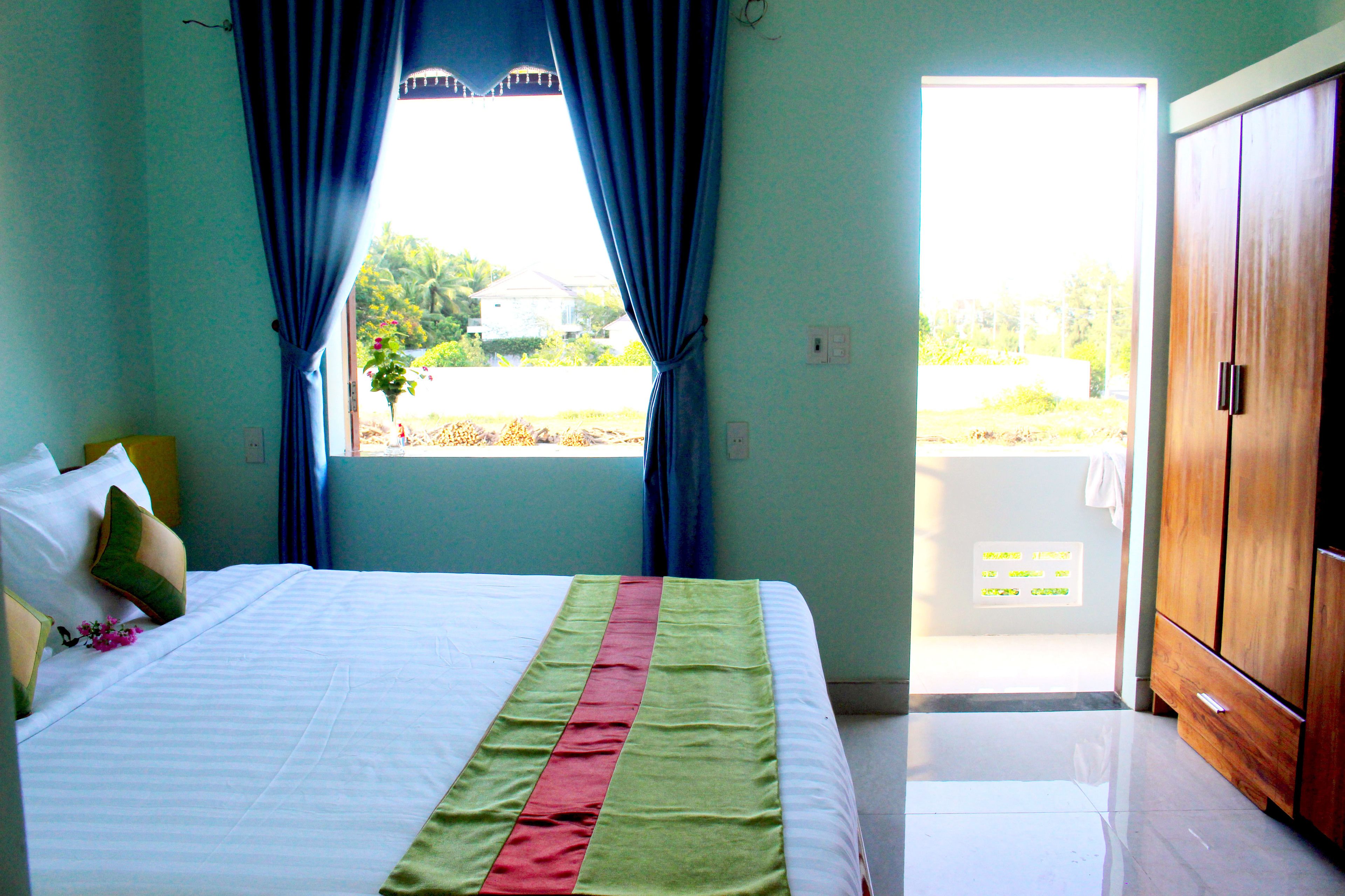 A bedroom with a bed, a window with curtains, a wooden wardrobe, and a view of greenery outside.