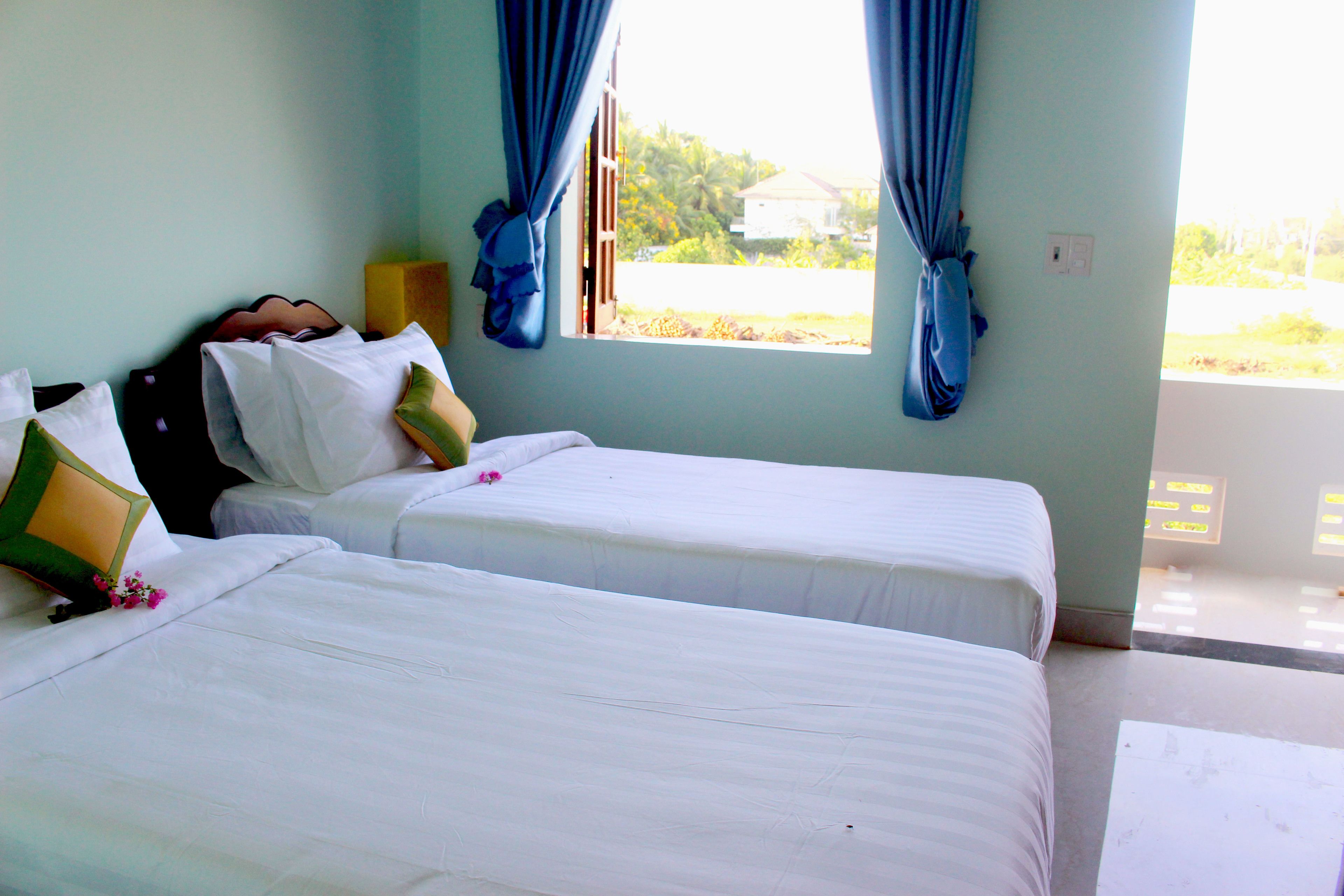 A room with two beds, a window with curtains, and a view of greenery outside.