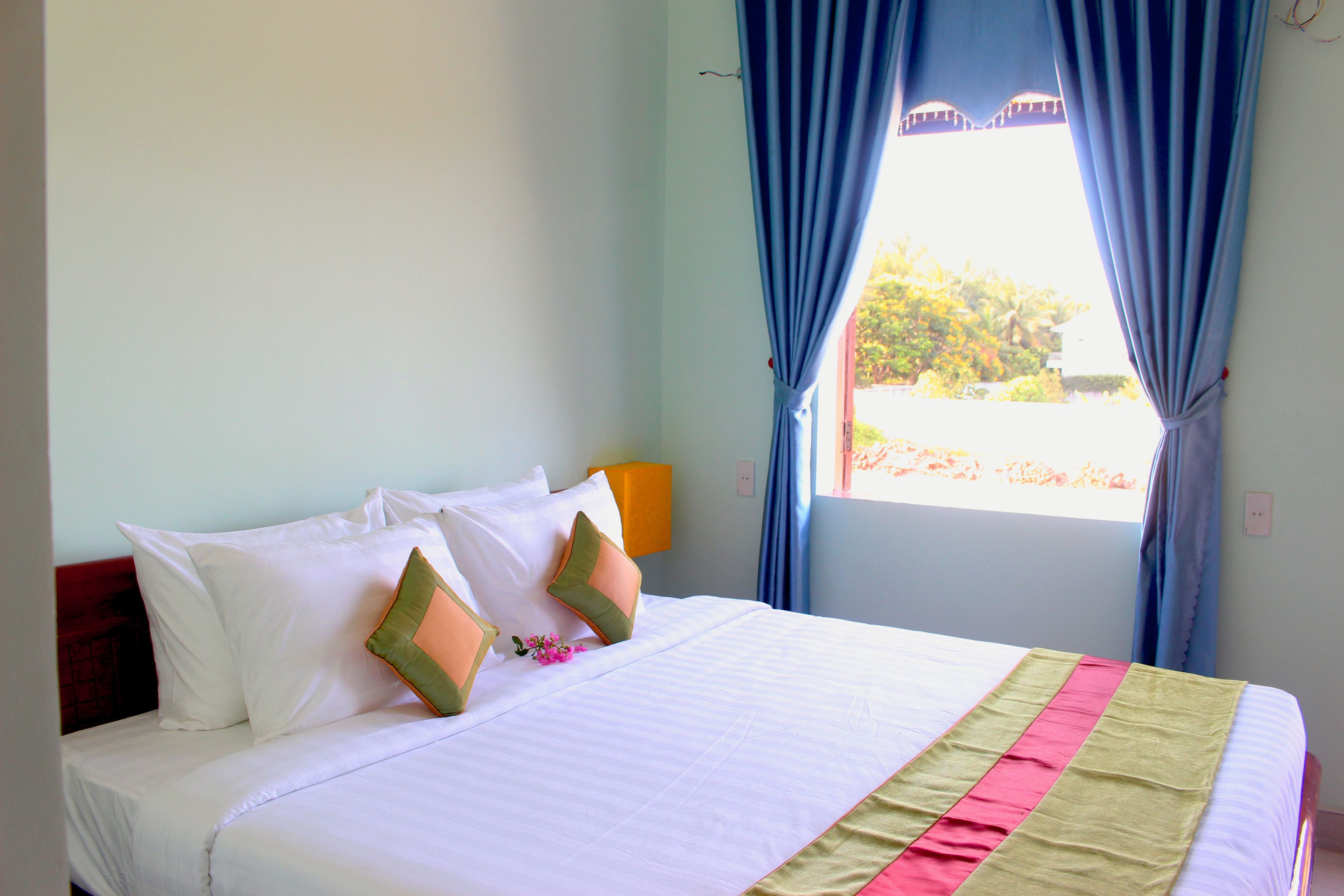 A neatly made bed with white linens and two green pillows, a window with blue curtains, and a wall-mounted lamp.