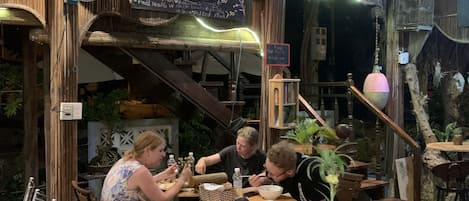 A rustic outdoor dining area with wooden tables and chairs, a chalkboard menu, and a large green vase on the table.