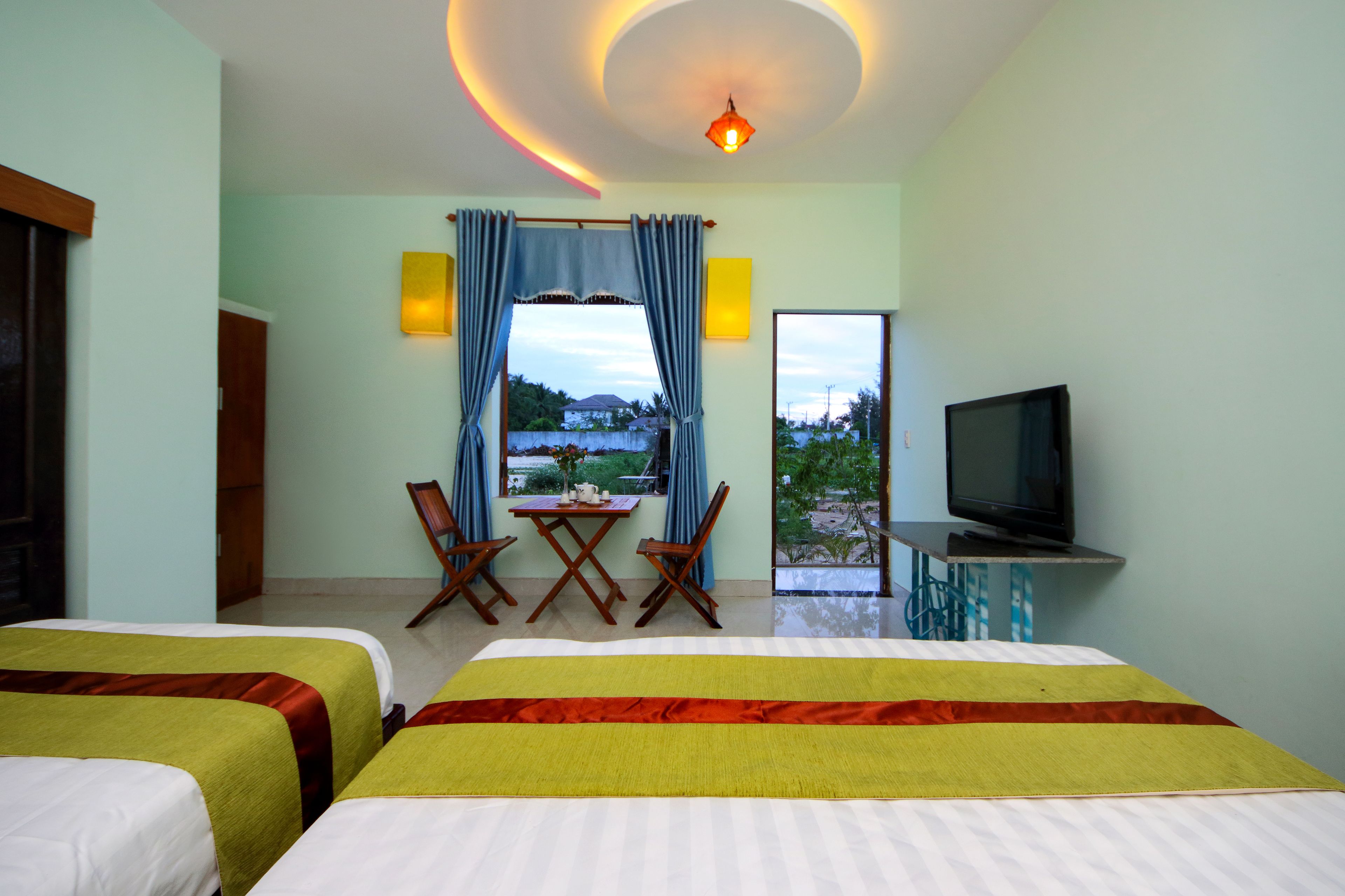 A hotel room with two beds, a TV, a dining table with chairs, and a view of the outdoors.