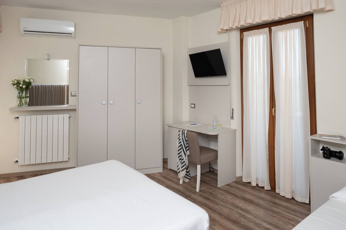 A hotel room with a bed, a desk with a chair, a TV mounted on the wall, a wardrobe, and a window with curtains.