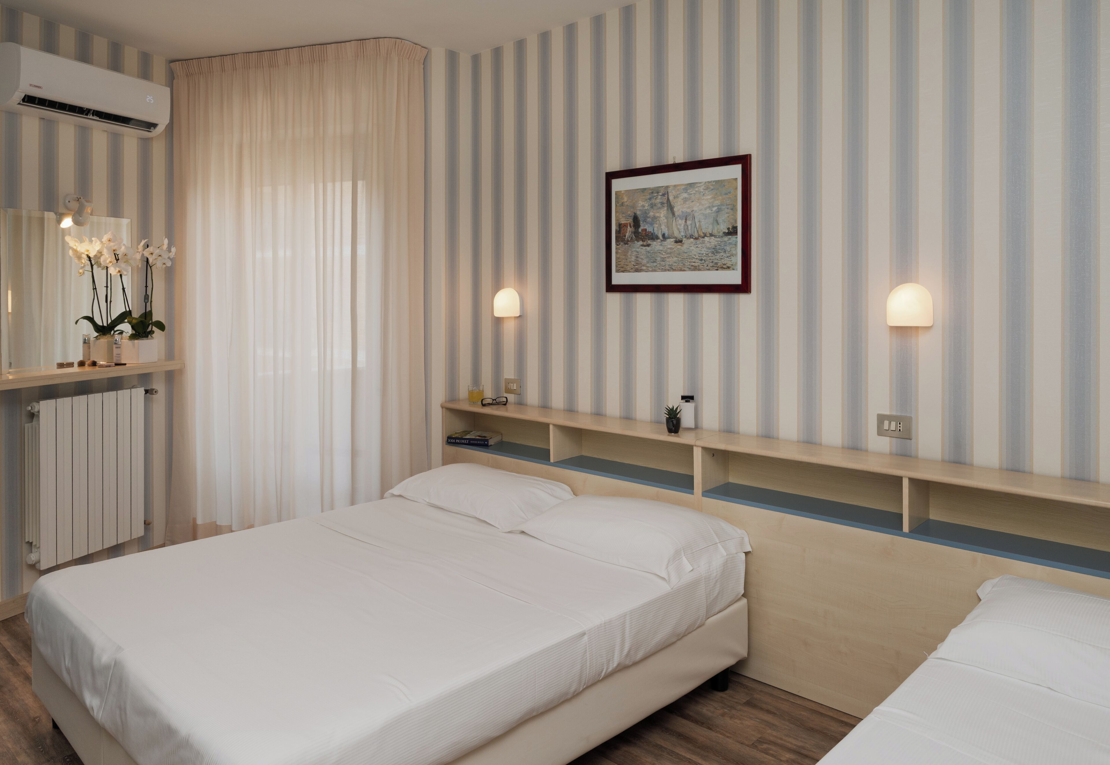 A hotel room with a bed, bedside tables, a mirror, an air conditioner, and a painting on the wall.