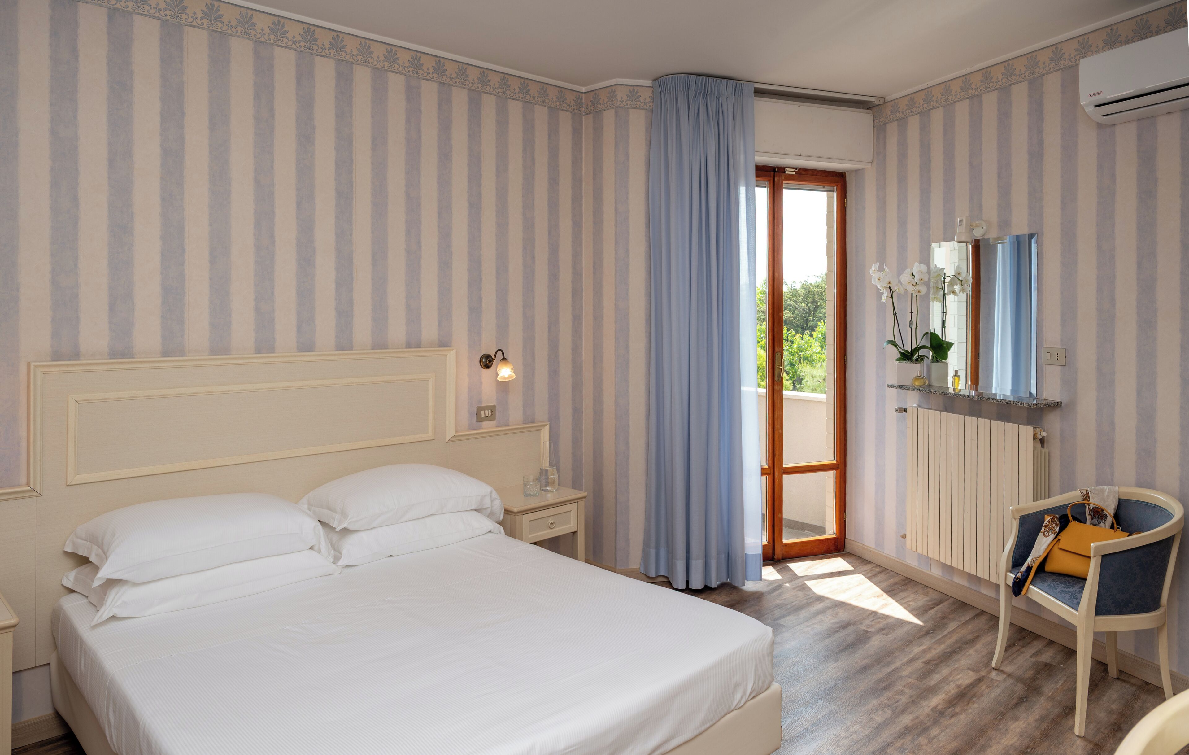 A hotel room with a large bed, a nightstand, a chair, a window with curtains, and a radiator.
