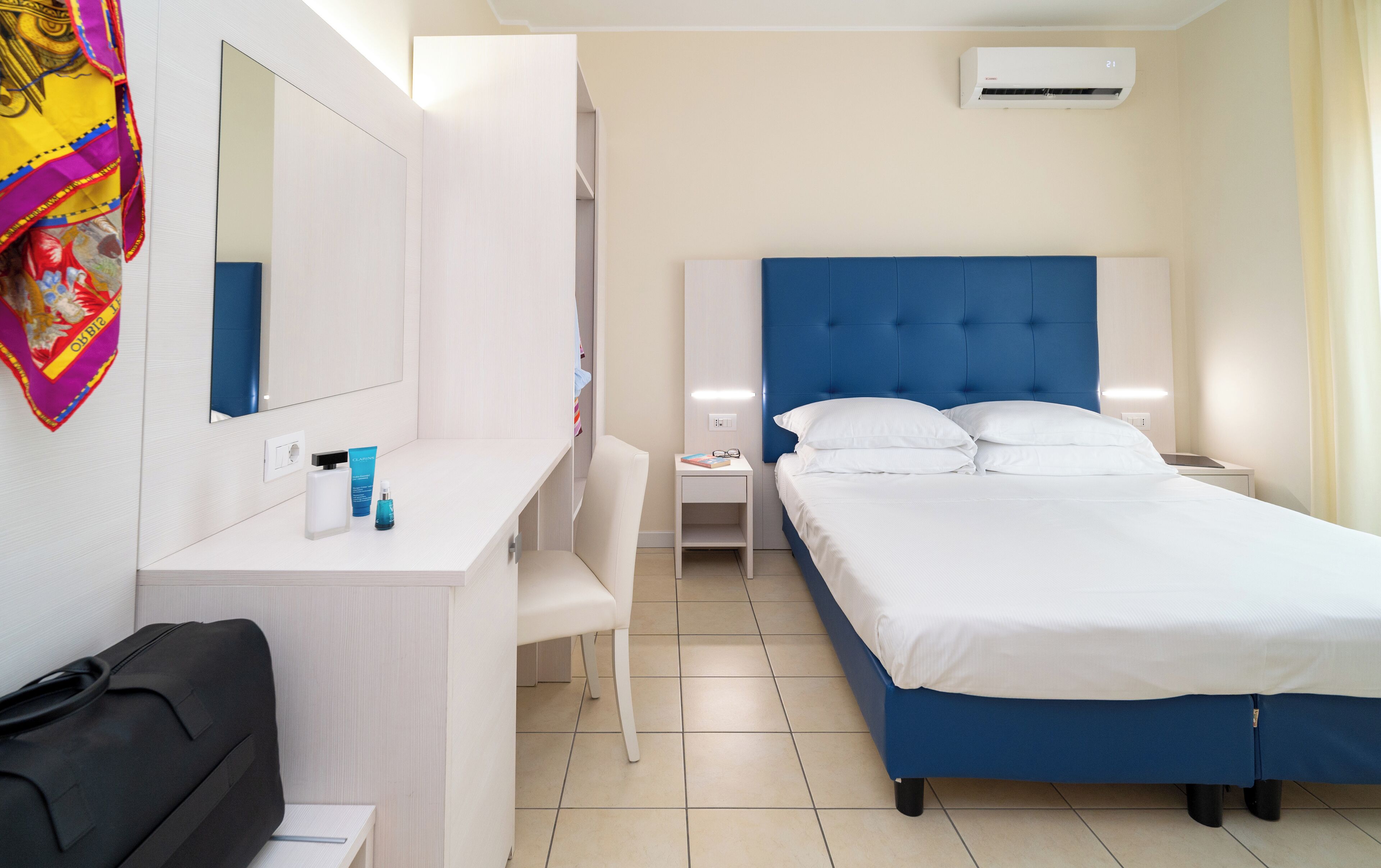 Quarto de hotel com cama, secretária, cadeira, mala, espelho e ar condicionado.
