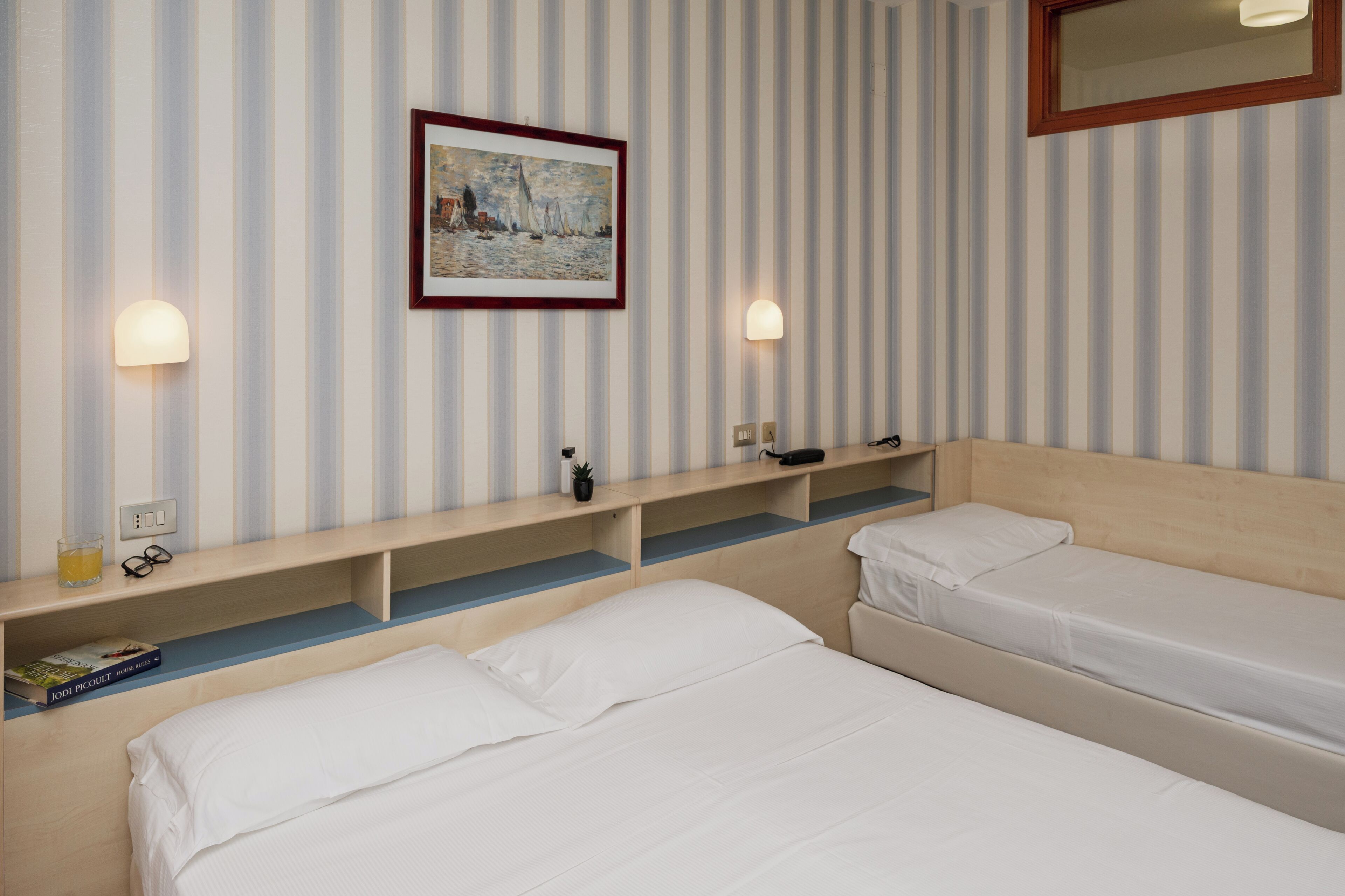 A hotel room with two single beds, a bedside table with a lamp, a framed painting on the wall, and a wooden shelf with a remote control.