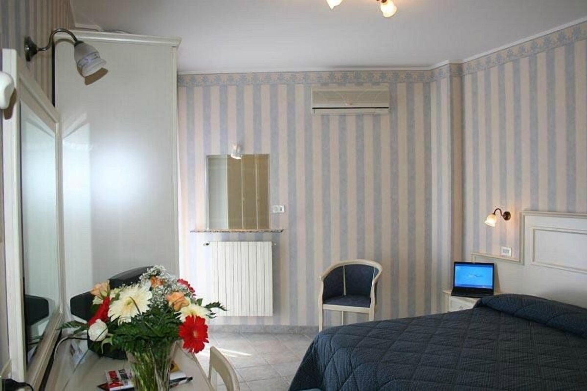 A hotel room with a bed, a desk with a computer, a chair, a mirror, and a radiator.