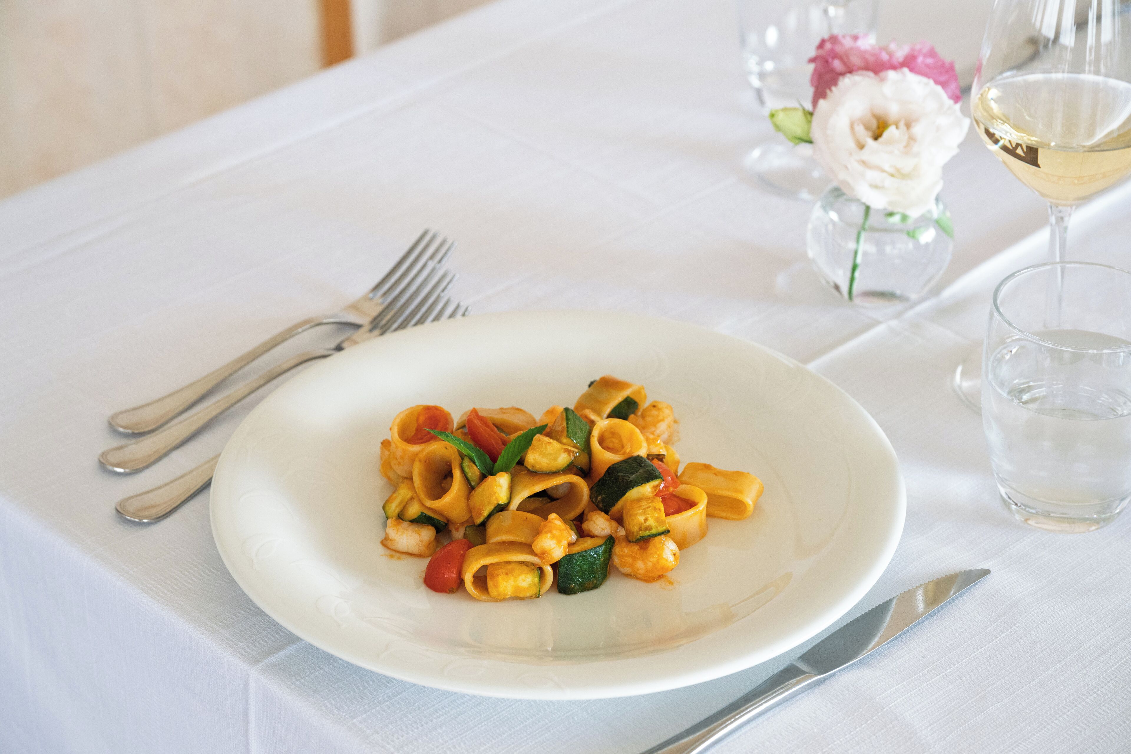 A white plate of pasta with vegetables and shrimp, a fork, a knife, a glass of water, and a glass of white wine on a white tablecloth.