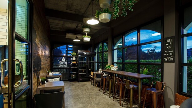 A cozy cafe with a rustic interior, featuring a long wooden bar, multiple seating options, and large windows offering views of the outdoors. - Sata House Buoutique Resort (Nakhon Ratchasima)