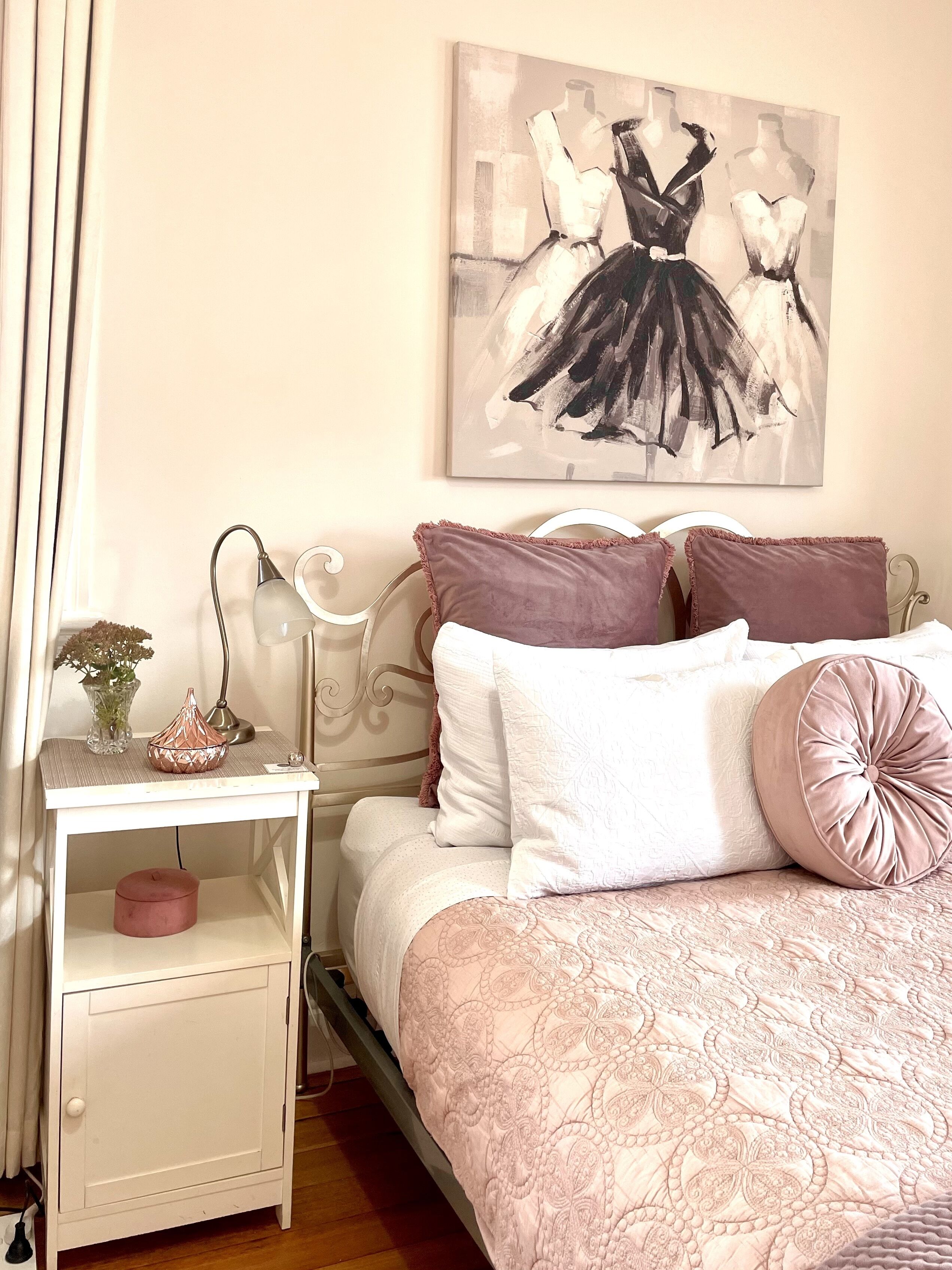 A bedroom with a bed, nightstand, lamp, and a painting of ballerinas.