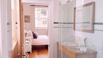 Un pasillo estrecho que da a un dormitorio con cama y ventana, un baño con mampara de vidrio, lavabo y espejo, y un banco de madera.