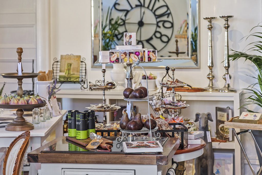 Una presentación ordenada de alimentos y bebidas, que incluye varios quesos, frutas y productos envasados, dispuestos en un expositor de varios niveles en una habitación con un reloj y elementos decorativos al fondo.